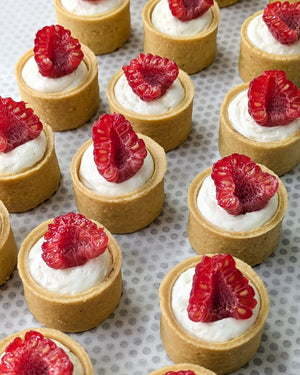 A batch of mini raspberry tarts with a white cream filling and a raspberry on top, arranged on a surface with a gridded pattern.
