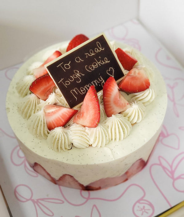 A strawberry cake with two layers, buttery cake with vanilla cream and fresh strawberries, with a decorative topper that reads 'To a real tough cookie, Happy Birthday!' on a pink floral background.